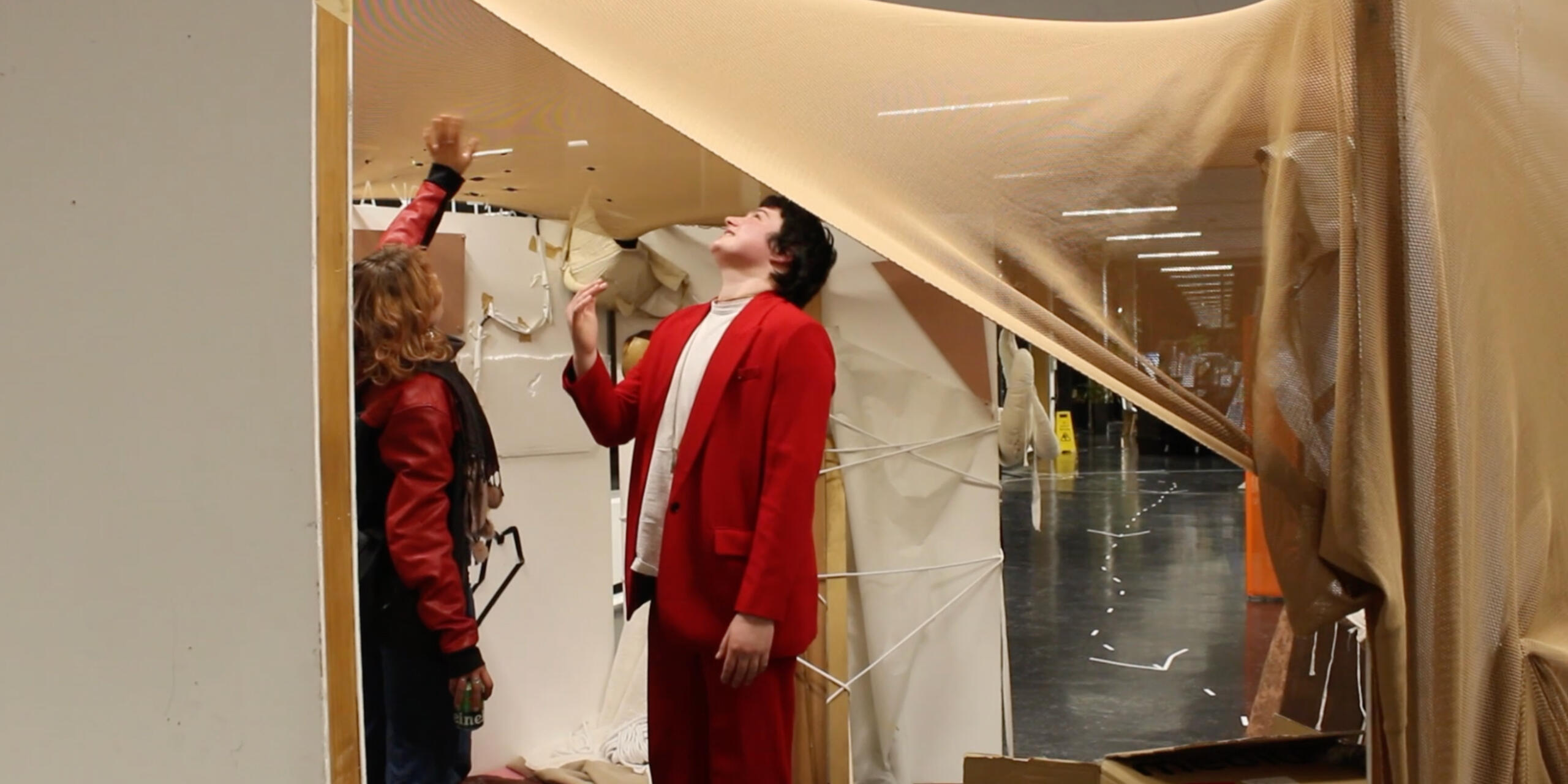 two people wearing red jackets look up at the textile that forms the ceiling of a booth, a sound art and set design installation in Deptford, 2023.